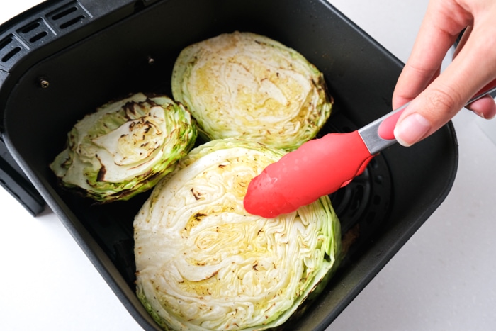 red tipped tongs flipping cooked cabbage steak out of air fryer tray on counter.