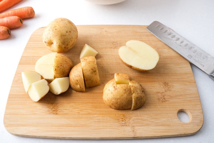 potatoes cut into smaller cubes on wooden cutting board with knife beside on white counter.