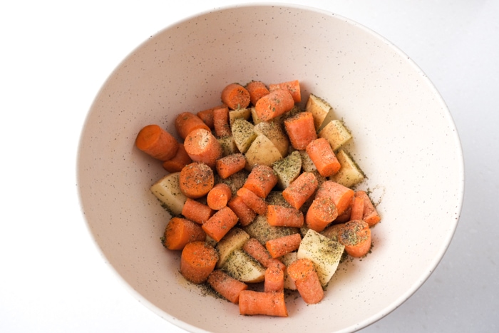 cut raw potatoes and carrots in white mixing bowl with oil and spices on them.