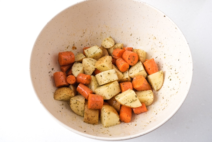 cut potatoes and carrots in white bowl covered in oil and spices sitting on white counter.