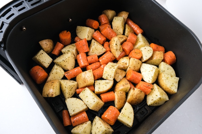 raw potatoes and carrots covered in oil and spices sitting in black air fryer tray on white counter.