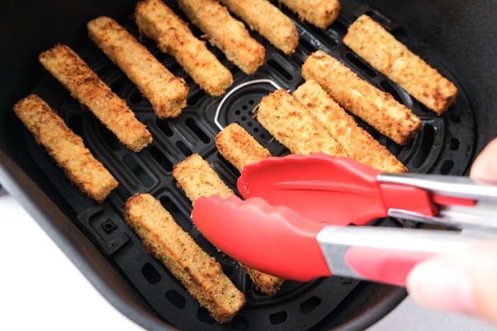 red tipped tongs flipping breaded eggplant fries sitting in black air fryer tray on counter.