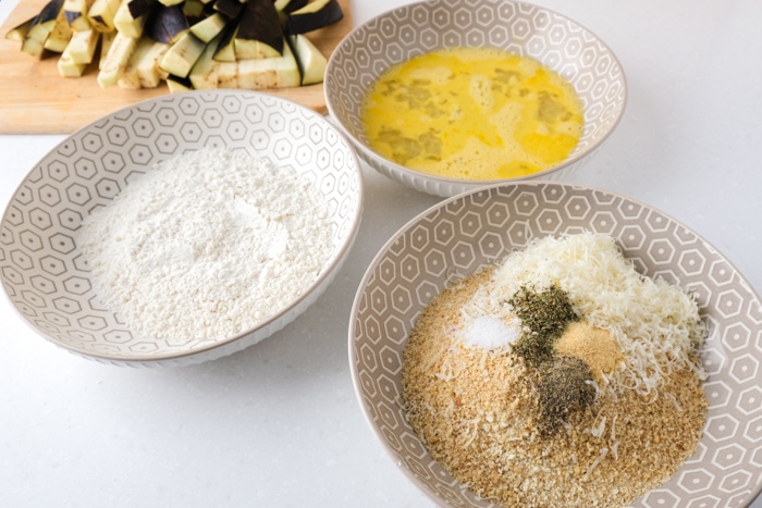 three bowls on white countertop filled with flour egg and breadcrumbs.