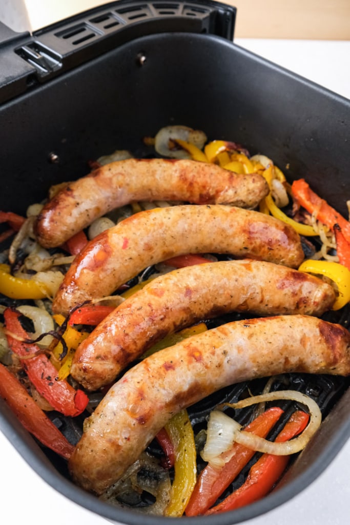 four cooked italian sausage sitting on cooked peppers and onions in black air fryer tray on counter top.