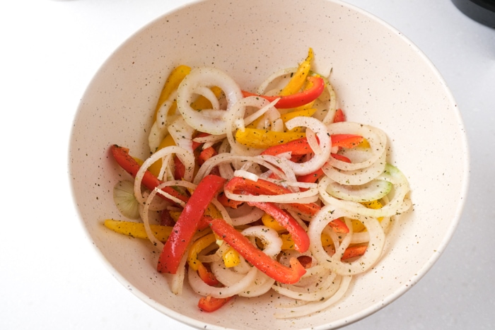 raw sliced onions and peppers covered in spices in white mixing bowl sitting on white counter.