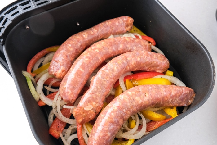 raw italian sausages sitting on raw peppers and onions in black air fryer tray on counter.