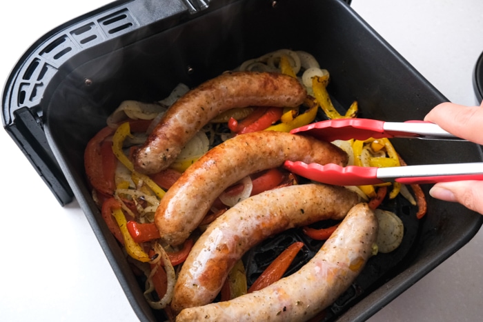 red tongs flipping cooked italian sausage sitting on peppers and onions in black air fryer tray.