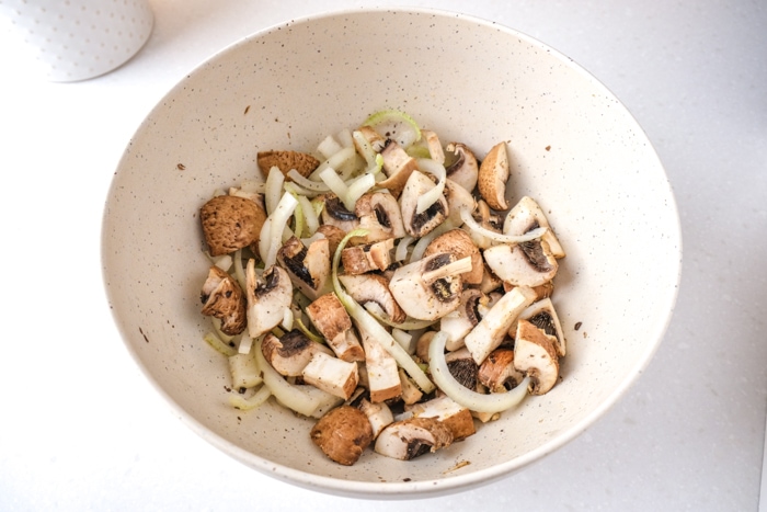 slices onions and cut mushrooms coated in oil and spices in white mixing bowl on counter top.