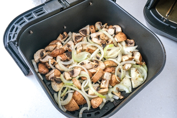 raw onions and mushrooms pieces in black air fryer tray on white counter.