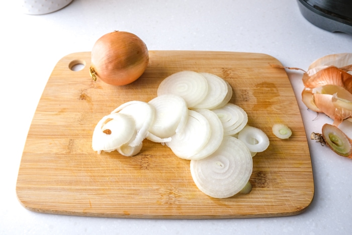 slices of white onions on wooden cutting board with whole onion behind on counter.