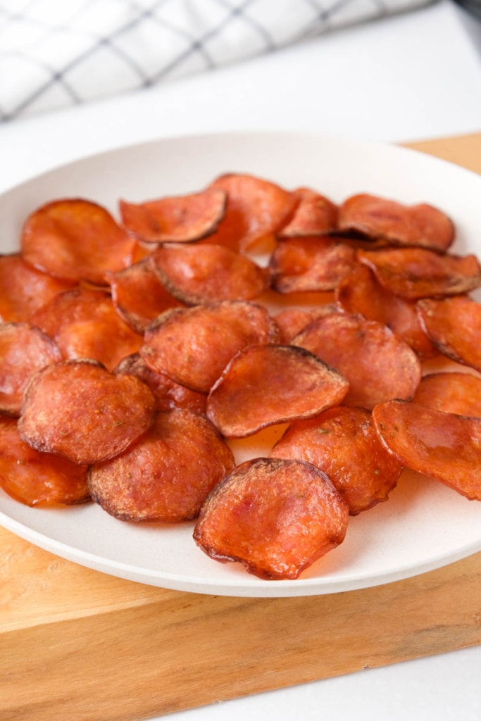 white plate of pepperoni chips on wooden board with white towel behind.