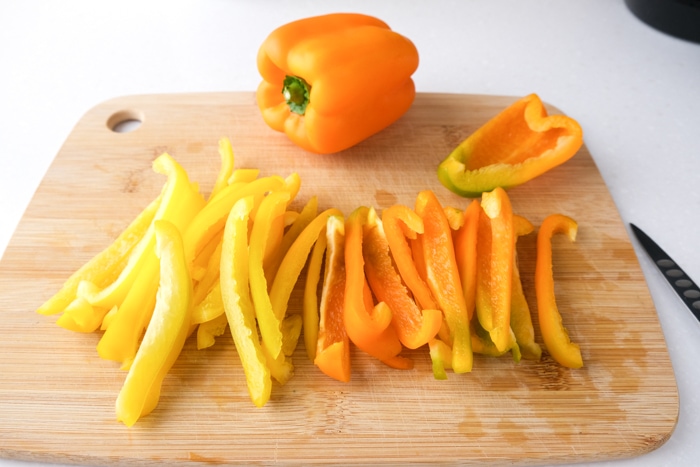 orange bell pepper on wooden cutting board with pepper strips cut up in front.