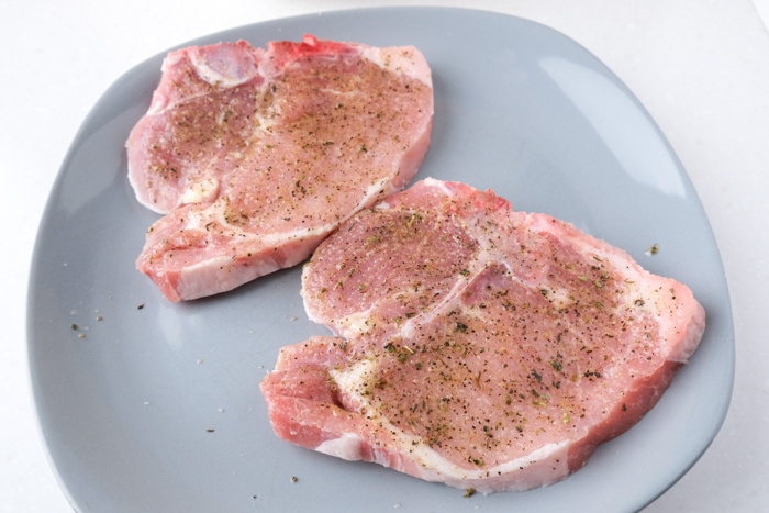 two raw pork chops sitting on blue plate with spices rubbed on on white countertop.