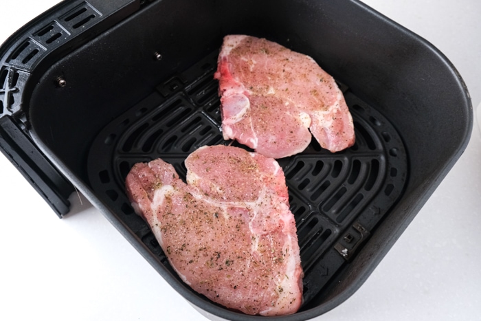 two raw pork chops with spices on top sitting in black air fryer tray.
