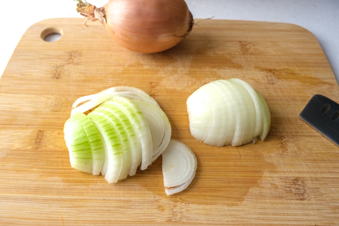 whole onion on wooden cutting board behind half a peeled onion cut into slices also on cutting board.