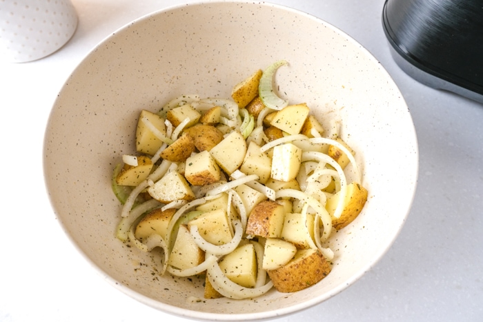 raw onions and potatoes coated in oil and spices in white mixing bowl on white counter with air fryer behind.