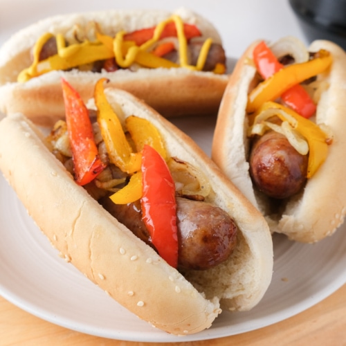 three brats in buns with peppers on top on white plate sitting on wooden board.