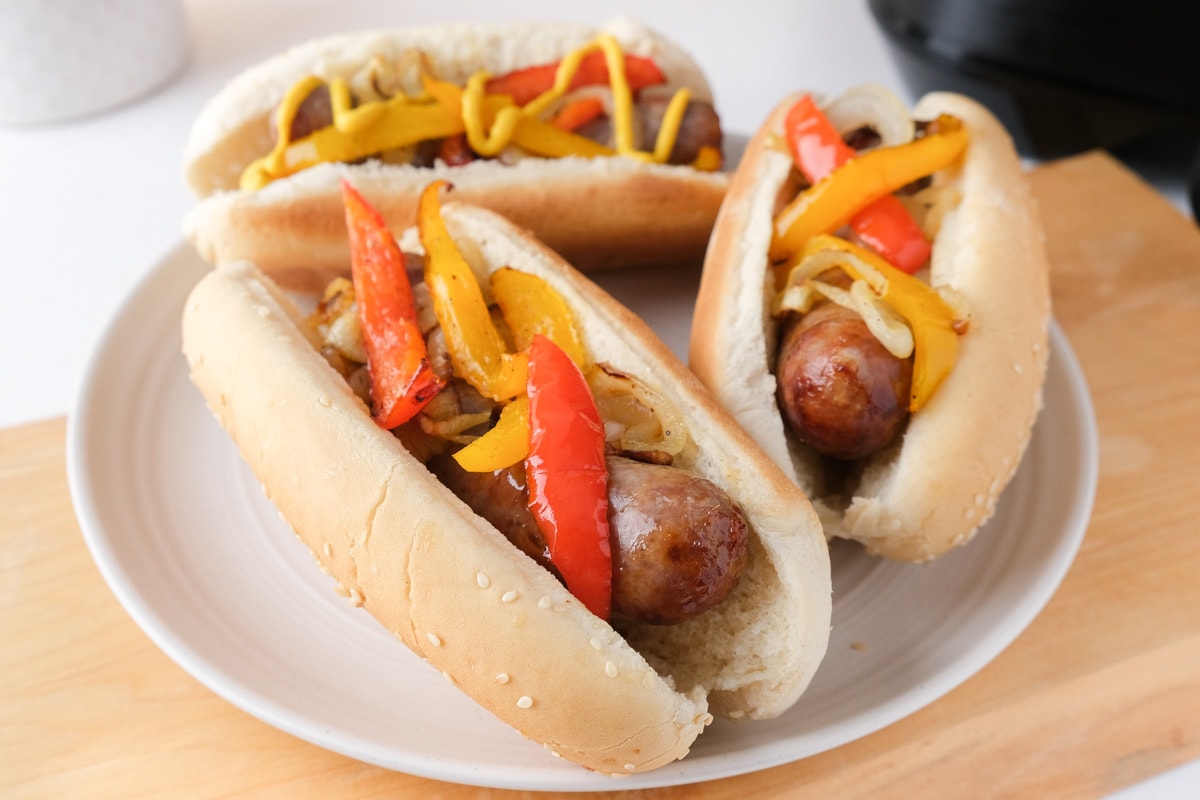 three brats in buns with peppers on top on white plate sitting on wooden board.