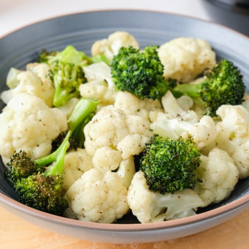 blue bowl filled with cooked cauliflower and broccoli florets sitting on wooden board.