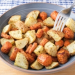 blue bowl of cooked carrots and potatoes with silver fork sticking out on wooden board.
