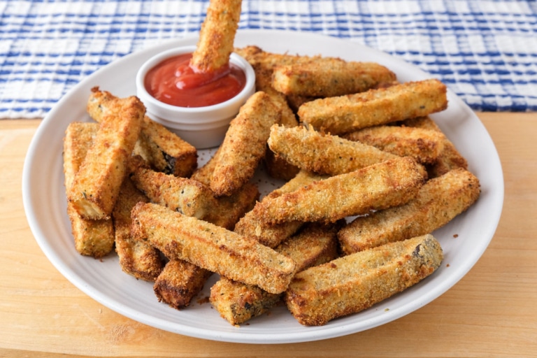 breaded eggplant fries on white plate sitting on wooden board with one fry dipping in ketchup.