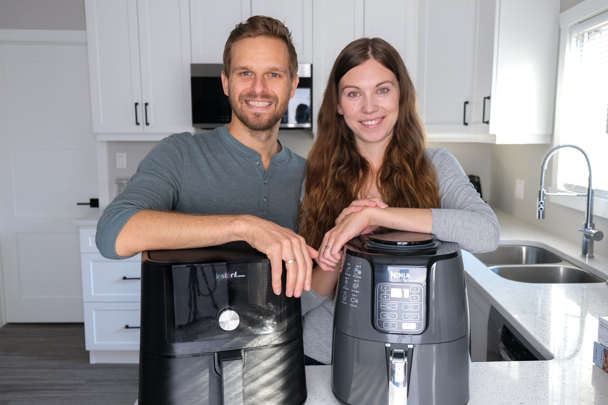 man and woman standing behind two black air fryers in kitchen with white cupboards.