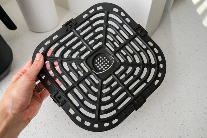 square black air fryer tray held in hand above white counter top in kitchen.