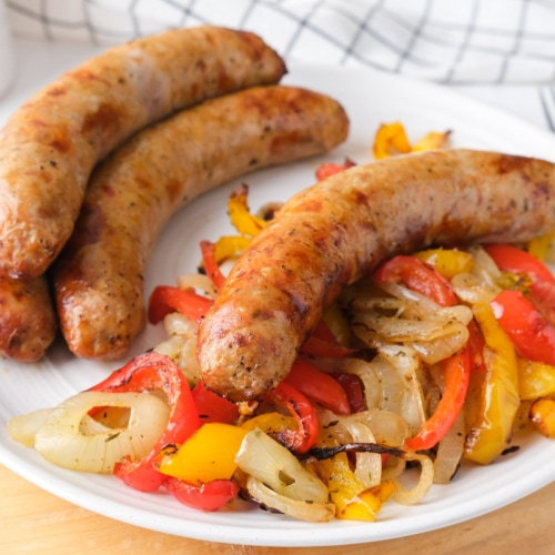 four Italian sausage on white plate with cooked peppers and onions sitting on wooden board on counter.