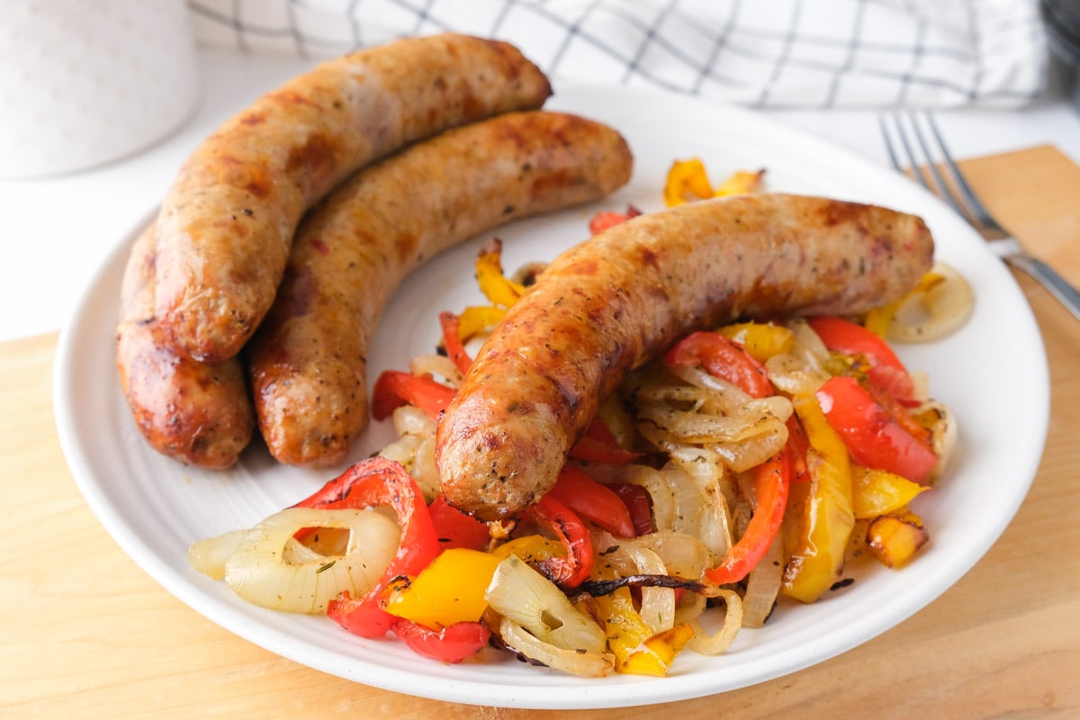four Italian sausage on white plate with cooked peppers and onions sitting on wooden board on counter.