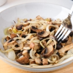 cooked mushrooms and onions in bowl on wooden board with silver fork sticking out.