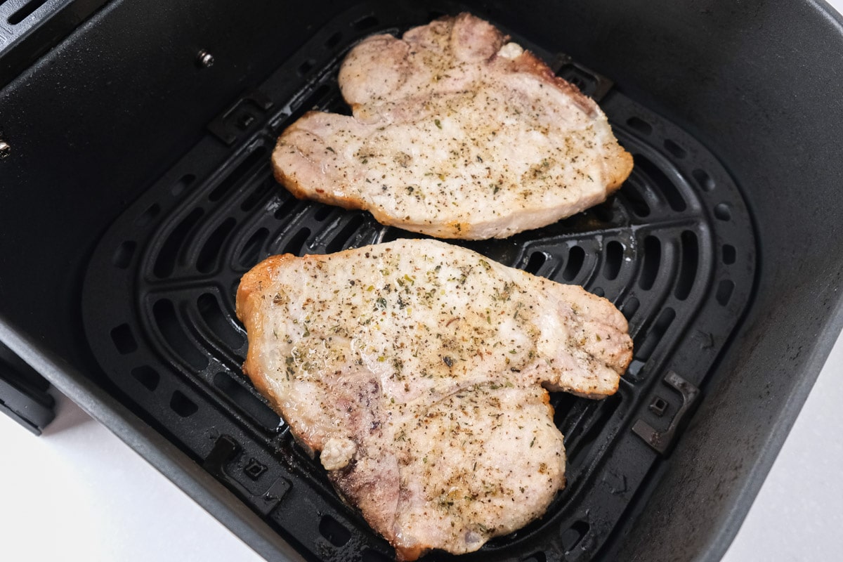 two cooked pork chops in black air fryer basket on white countertop.
