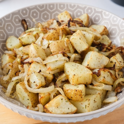 cooked potatoes and onions in bowl sitting on wooden board on counter with air fryer behind.