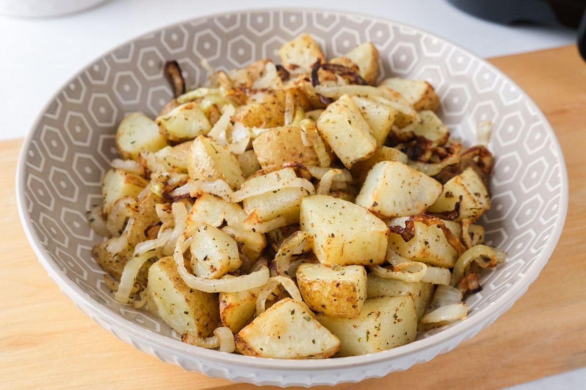 cooked potatoes and onions in bowl sitting on wooden board on counter with air fryer behind.
