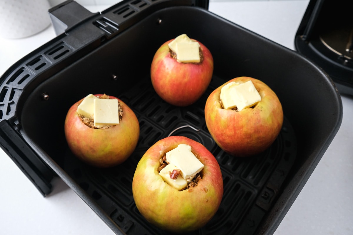 four apples with butter sitting on top sitting in black air fryer basket on counter.