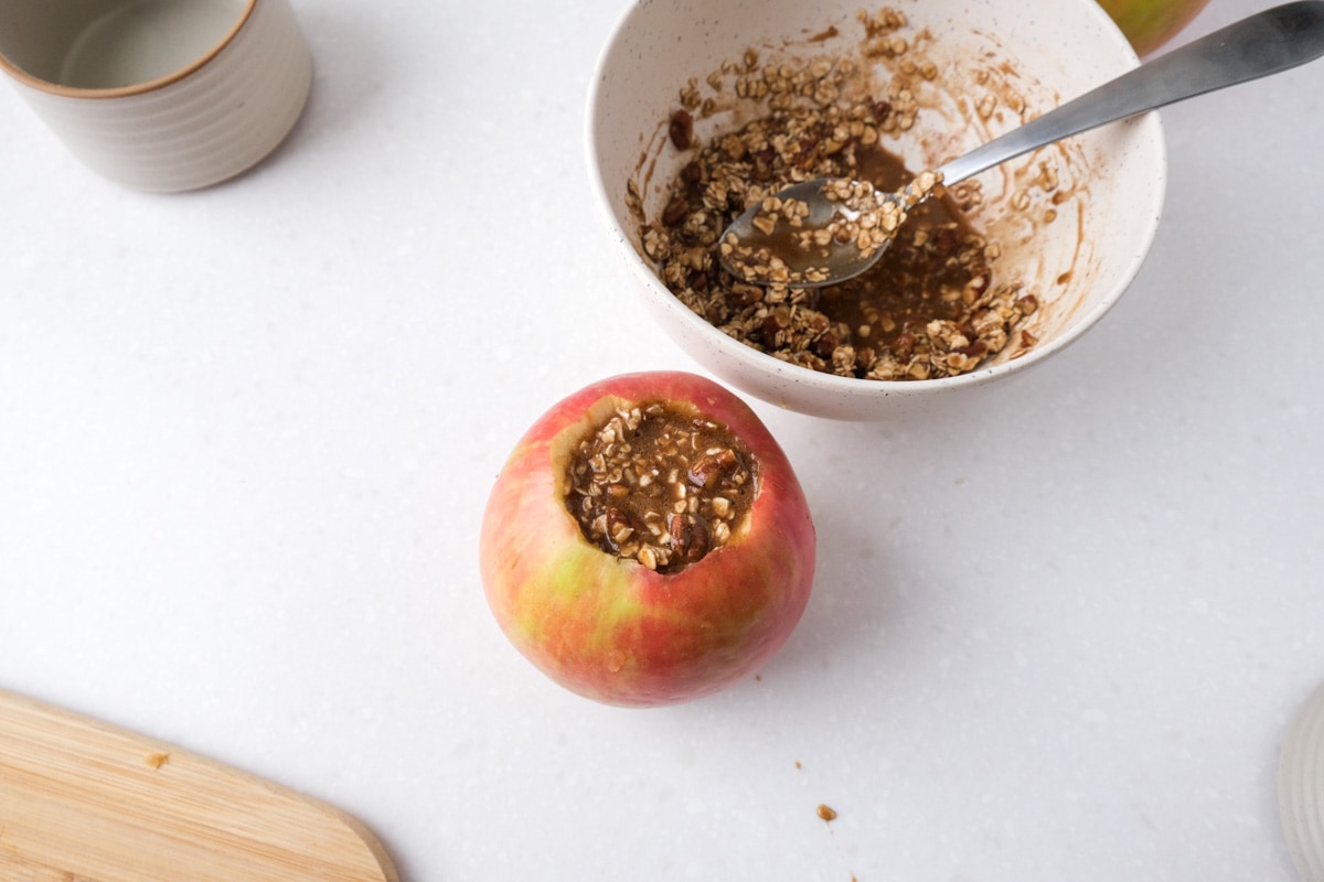 one cored apple filled with cinnamon oat mixture with bowl behind sitting on white counter.