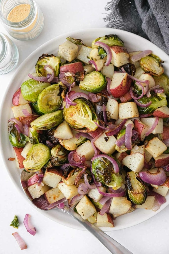 plate of brussels sprouts and potatoes with silver spoon on the edge sitting on white counter top.