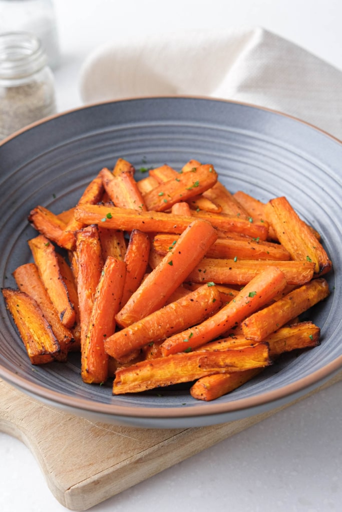 air fried carrot sticks in blue bowl on wooden board on white counter.