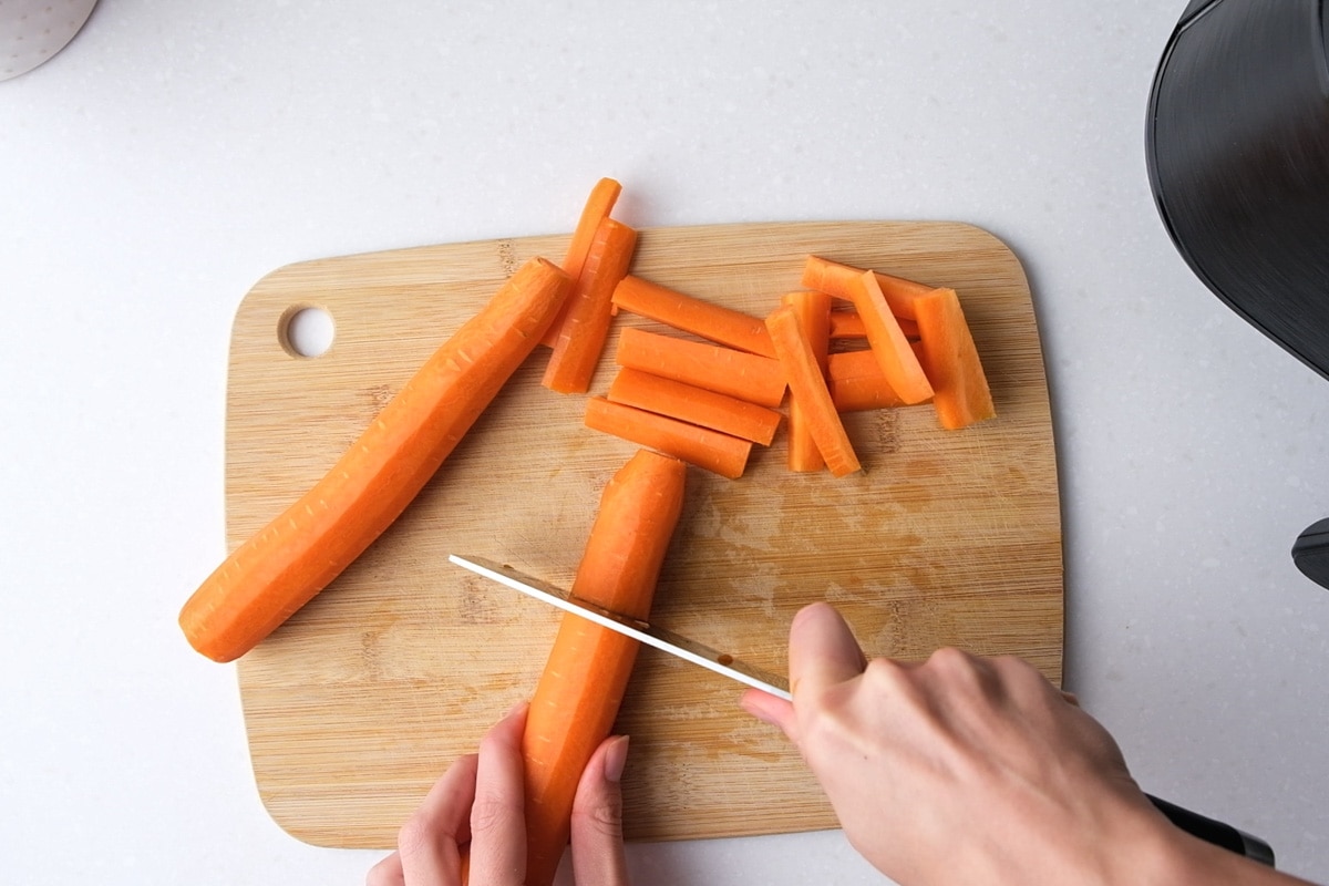 hand holding knife cutting carrots into chunks on wooden cutting board.