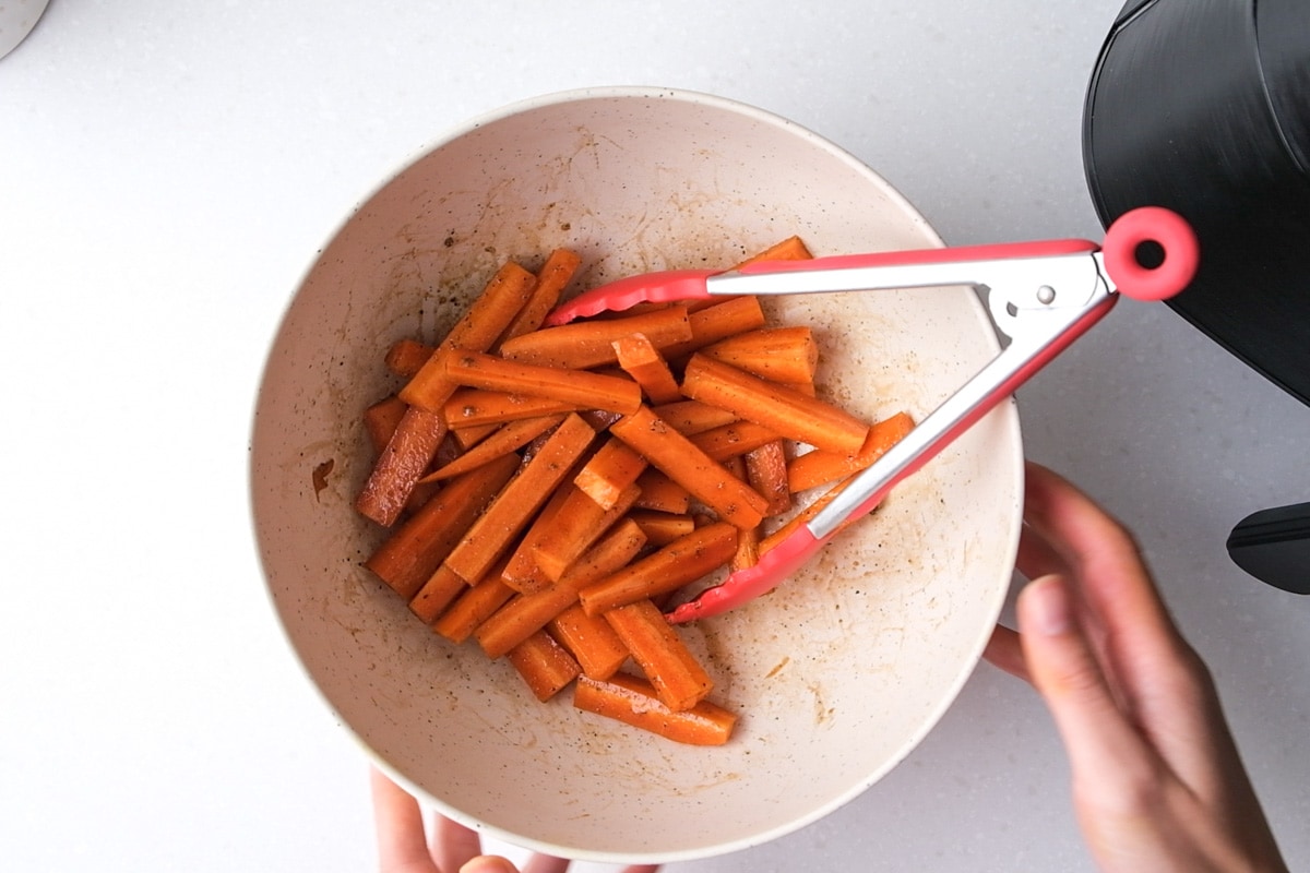 cut carrots coasted in oil and spices in white mixing bowl with red tipped tongs in the bowl.