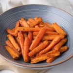 roasted carrots in blue bowl on wooden board with spices behind.