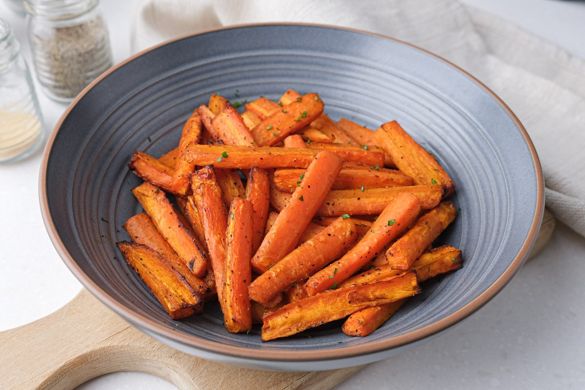 roasted carrots in blue bowl on wooden board with spices behind.