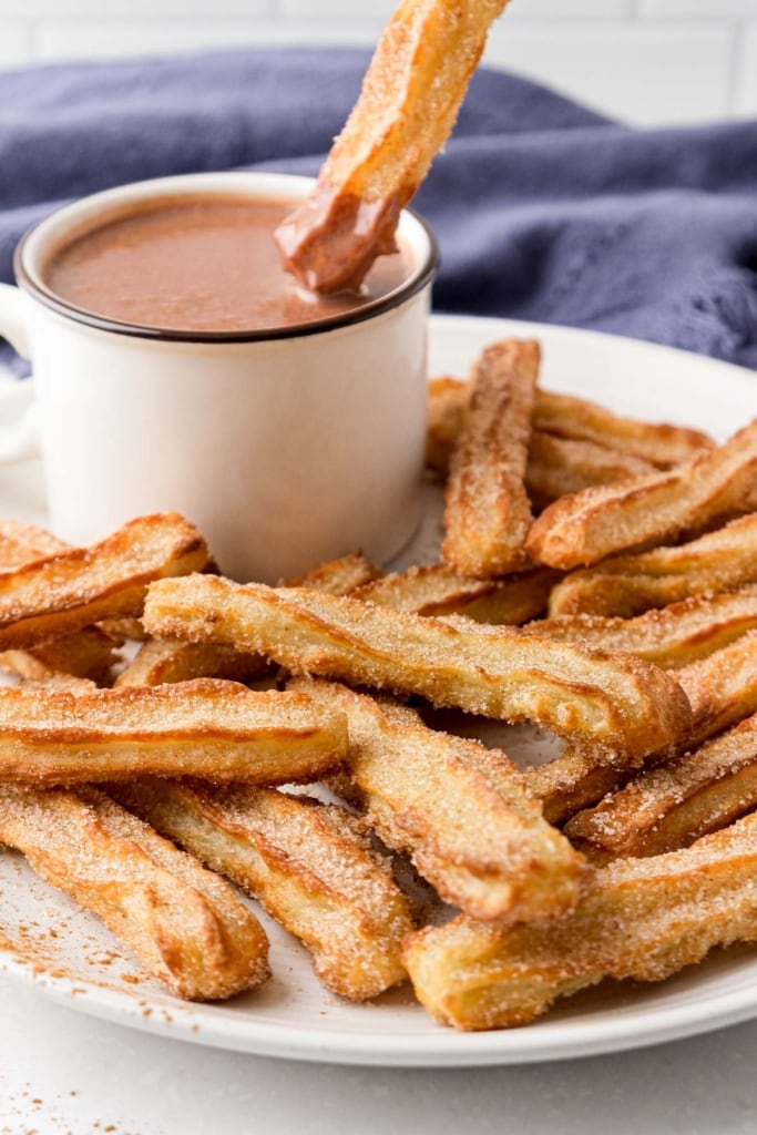 air fried churros on white plate with mug of hot chocolate behind with churros dipping into it.