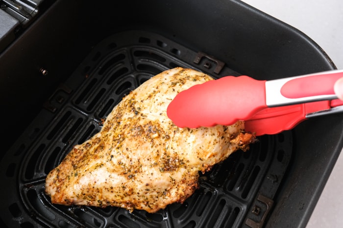red tipped tongs flipping coked turkey breast in black air fryer basket.