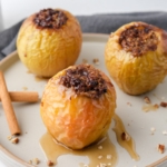three baked apples with oats on top on plate with cinnamon and syrup around.