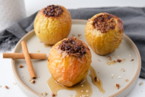 three baked apples with oats on top on plate with cinnamon and syrup around.
