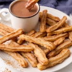 crispy churros on white plate with mug of hot chocolate behind.