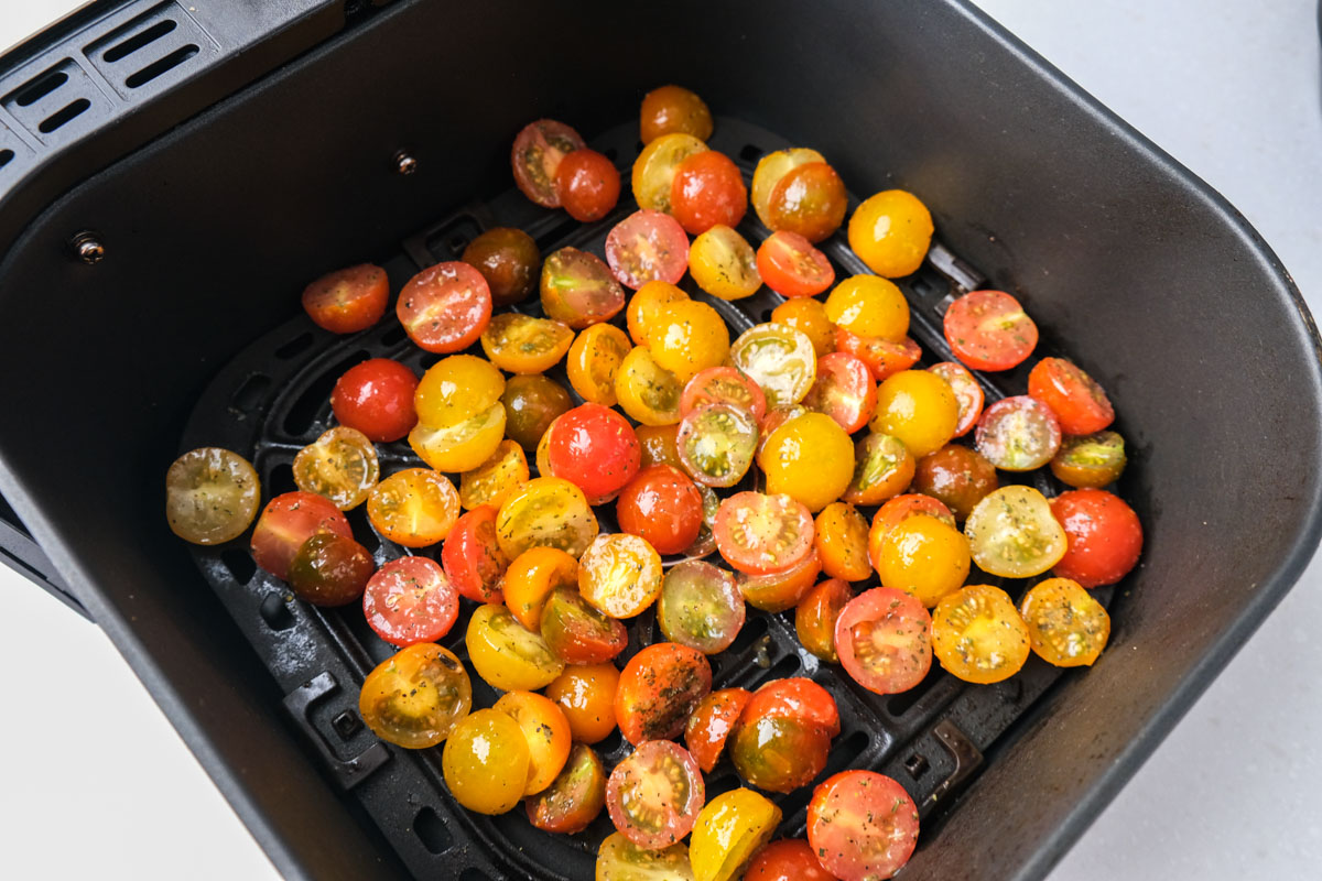 raw cherry tomatoes covered in oil and spices sitting in black air fryer tray on white counter.