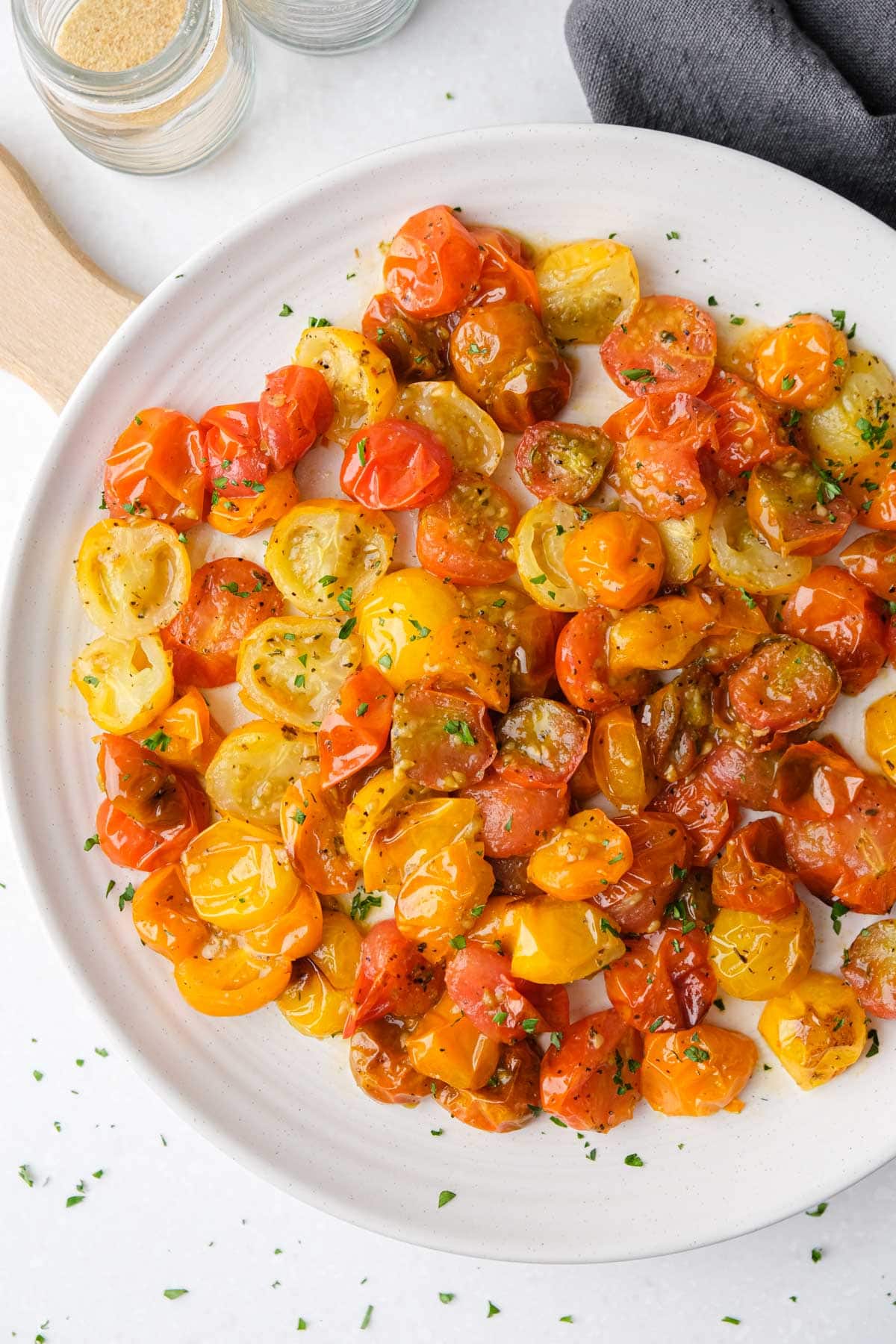 colorful roasted cherry tomatoes on white plate with wooden board and grey cloth beside.