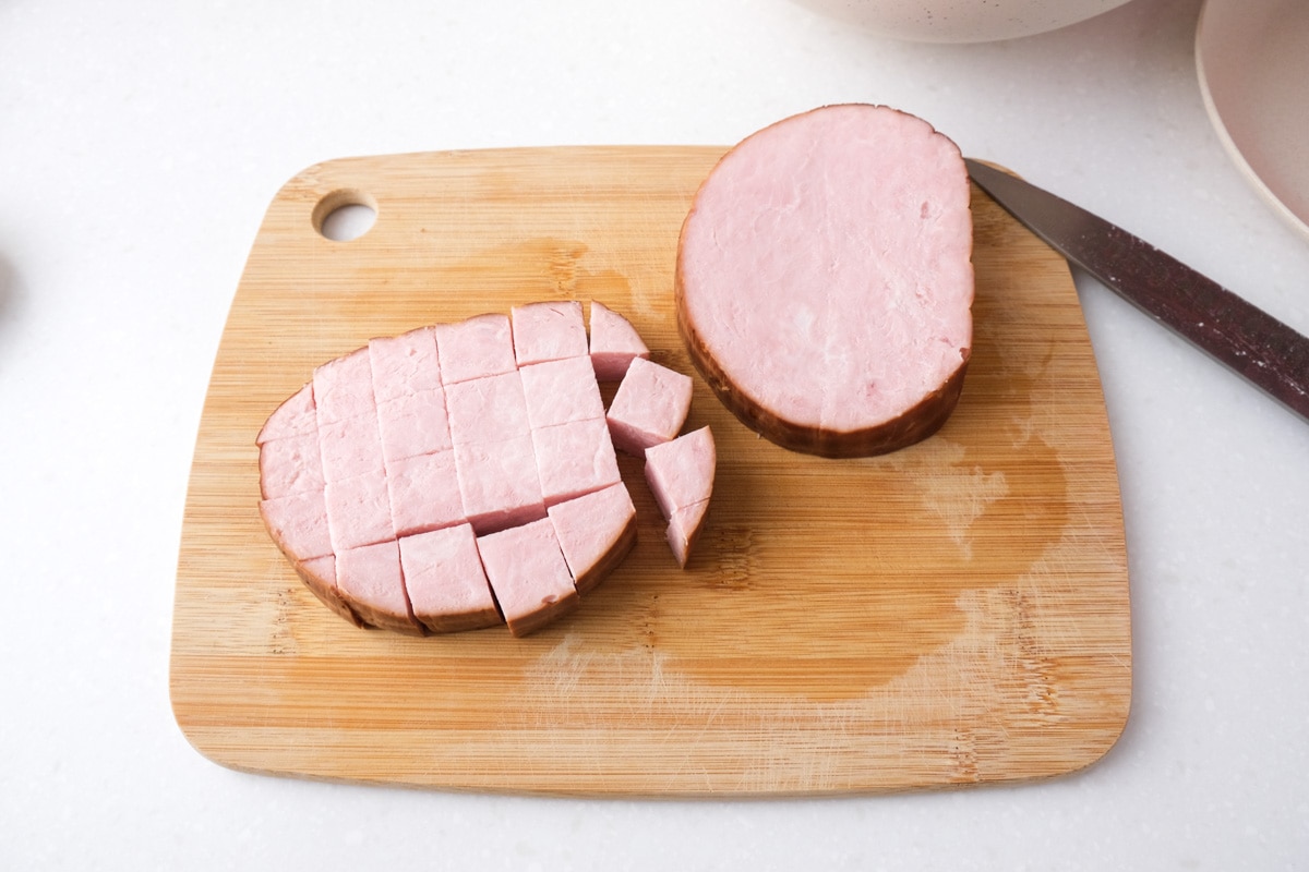 precooked ham cut into chunks with knife on wooden cutting board.