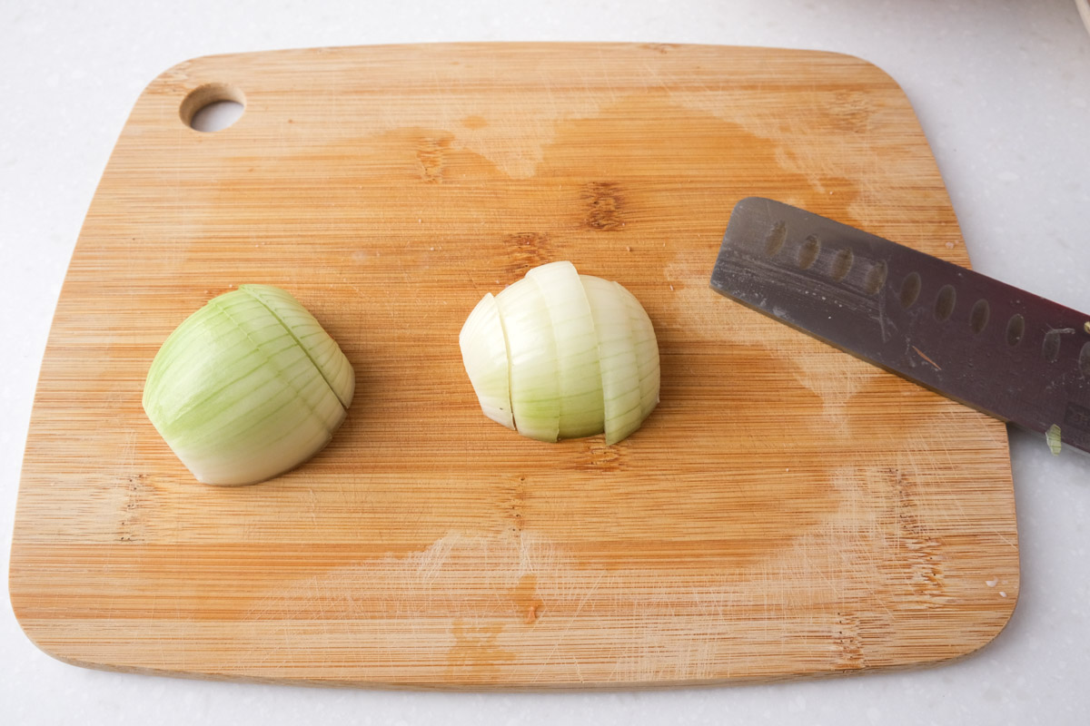 two halves of onion cut into slices on wooden cutting board with knife beside.